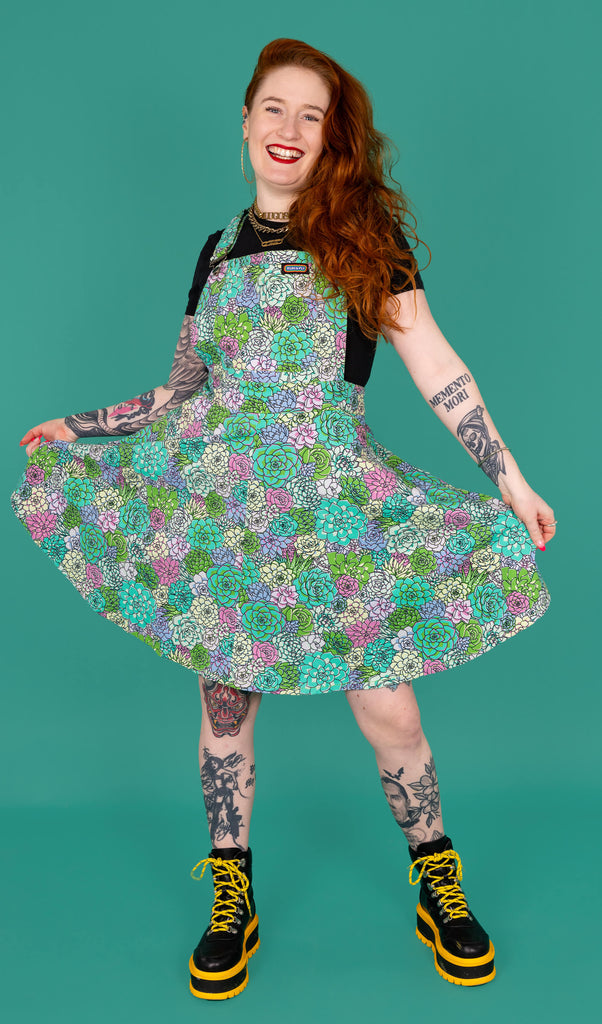 Isobella a tattooed model with long ginger hair is wearing a flared pinafore dress with mint green, blue and purple succulents all over paired with a black t-shirt and black and yellow boots. The model is smiling at the camera and swishing the skirt of the pinafore in front of a green background. 