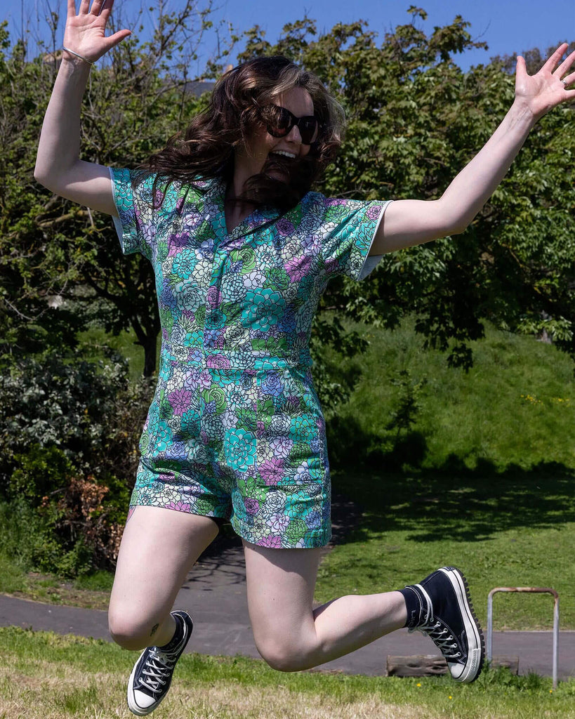 The Succulents Stretch Twill Playsuit worn by a femme model with long brown and blonde hair with black trainers and sunglasses. She is jumping in the air whilst smiling outside in a grassy sunny park area. The playsuit print features various sized succulents in pastel blue, pastel lilac, teal, pink and pale yellow.