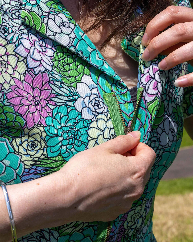 Close up of the Succulents Stretch Twill Playsuit worn by a femme model with long brown and blonde hair. She is outside in a grassy sunny park area showing the zip up front feature. The playsuit print features various sized succulents in pastel blue, pastel lilac, teal, pink and pale yellow.