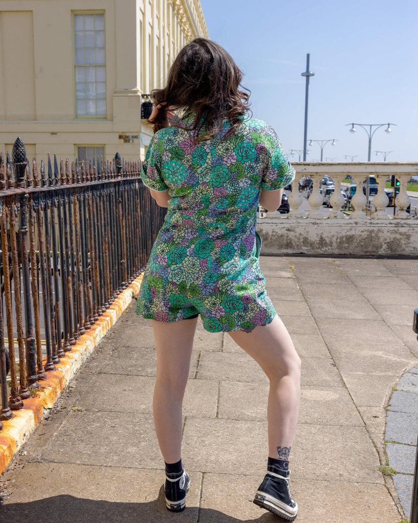The Succulents Stretch Twill Playsuit worn by a femme model with long brown and blonde hair. She is outside in a Hove facing away from camera. The playsuit print features various sized succulents in pastel blue, pastel lilac, teal, pink and pale yellow.