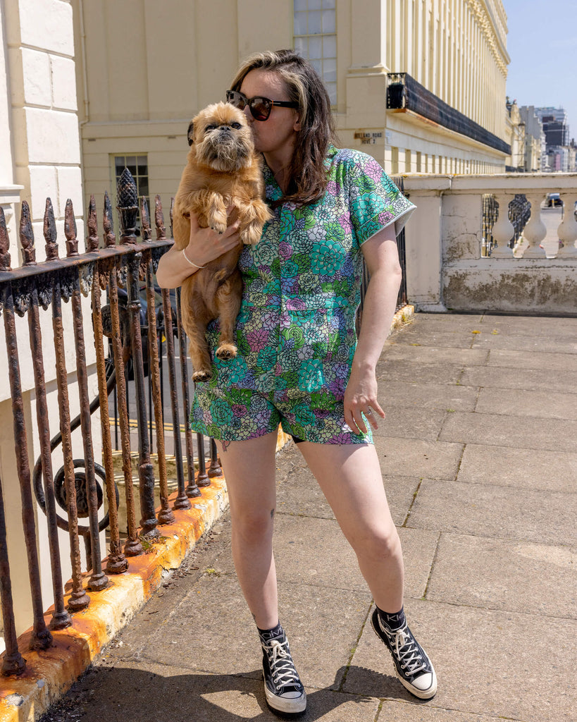 The Succulents Stretch Twill Playsuit worn by a femme model with long brown and blonde hair with black trainers and sunglasses. She is facing forward and holding Brian the dog. The playsuit print features various sized succulents in pastel blue, pastel lilac, teal, pink and pale yellow.