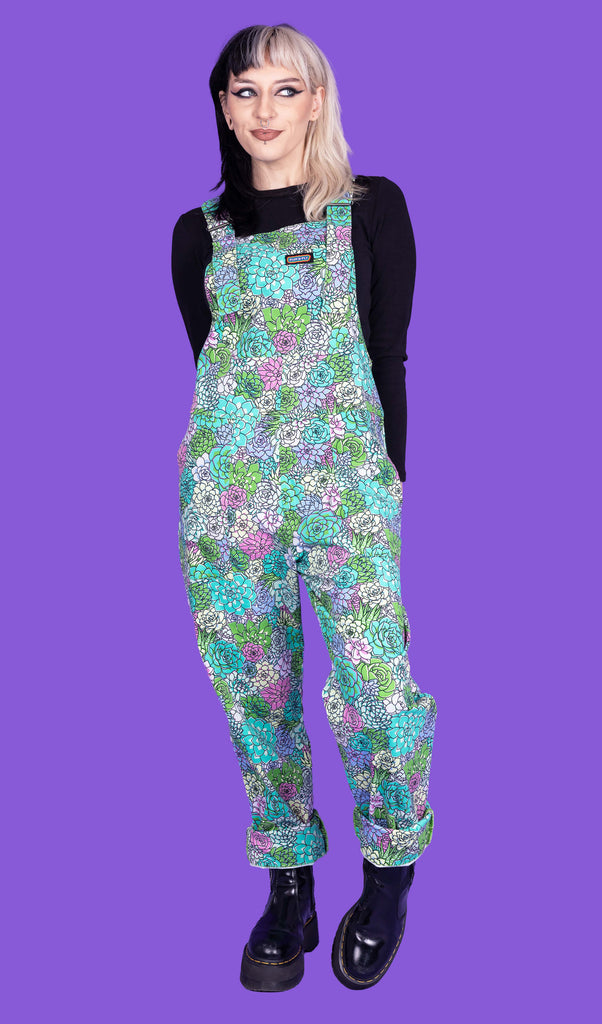 Claudia, a beautiful model with black and blonde split dyed hair is wearing Run & Fly dungarees with mint green, blue and purple succulents all over. The dungarees are paired with a long sleeved black top and black boots. Claudia is posing with one leg bent and hands behind her back in front of a purple background. 