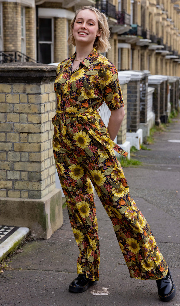 Amy is standing in front of a row of brick townhouses, wearing midnight blues sunflower jumpsuit in blue, green, yellow and orange with loose flared trouser legs, a waist tie, button up front with collared neck. Model is looking at the camera and smiling with arms behind back.