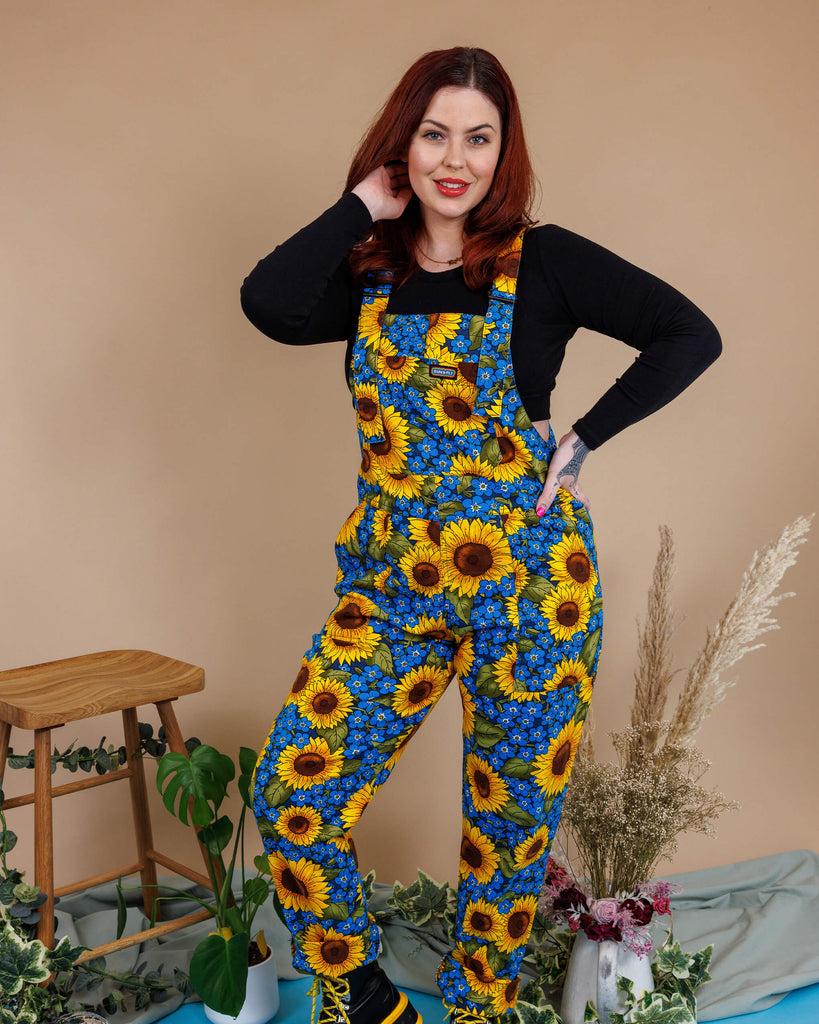 Aemelia Fox is stood in a photography studio in Hove with a tan colour backdrop and plants behind her wearing Run & Fly x Jen James Design Forget Me Not Charity Stretch Twill Dungarees with a long sleeve black top underneath and boots. The dungarees have an all over blue forget me not and yellow sunflower print. Aemelia is posing at the camera with one leg bent and one hand on her hip and other in her hair 