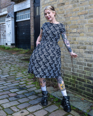 Tattooed model strikes a playful pose in the Run & Fly Swallow Print Dress. The monochrome bird pattern, waist tie, and pockets make it a stylish yet practical choice from this ethical slow fashion brand.