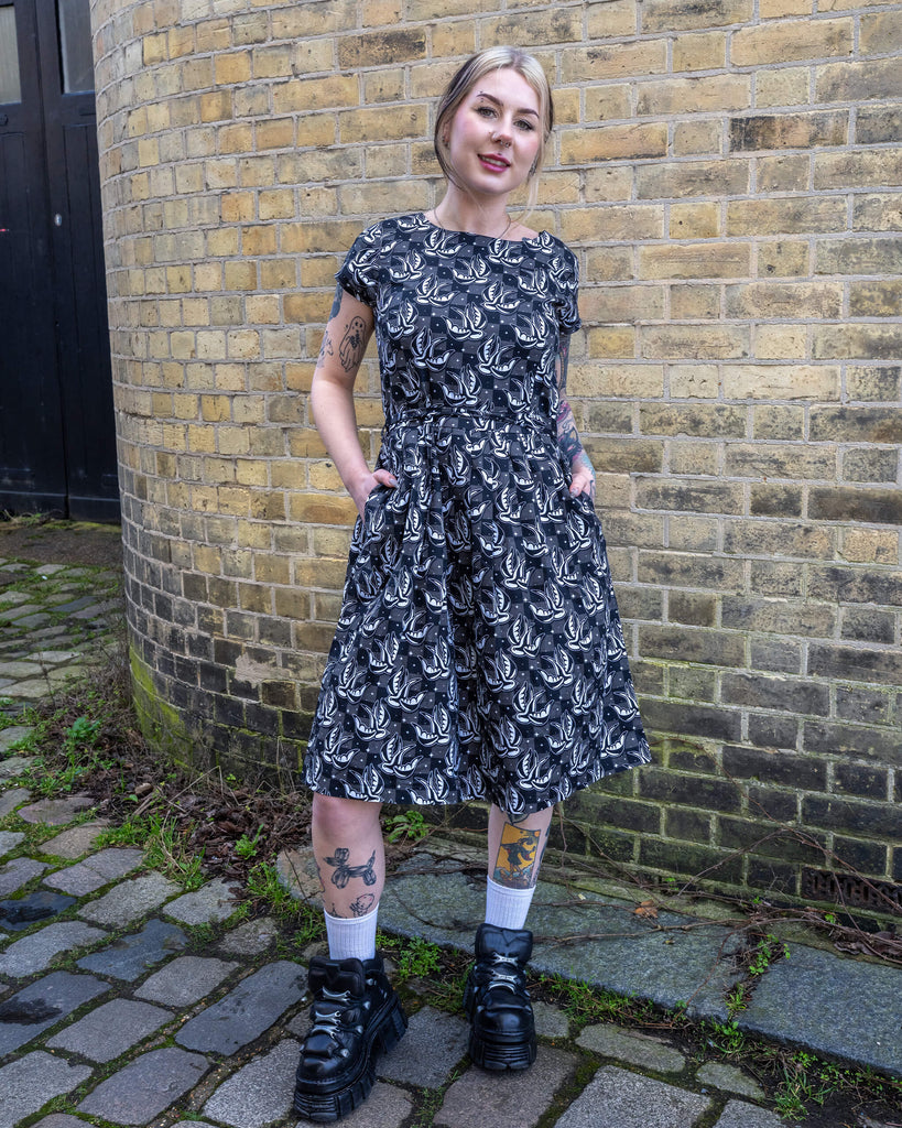Tattooed model smiling while wearing the Run & Fly Swallow Print Dress. The fit-and-flare design, tie waist, and pockets make it a standout piece for everyday wear. Ethically made for conscious fashion lovers.