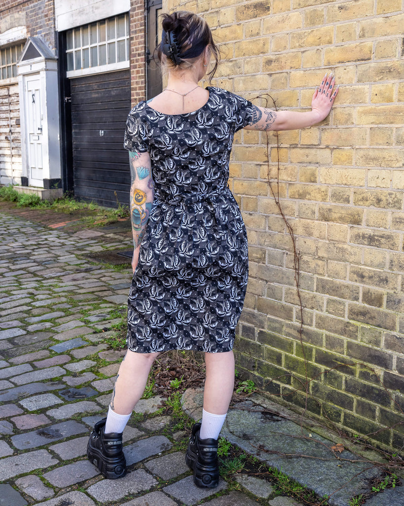 Tattooed model poses against a brick wall, wearing the Run & Fly Swallow Print Dress. The black and white bird design pops against the checkered background, and the tie-waist detail adds shape. Ethical slow fashion at its finest.