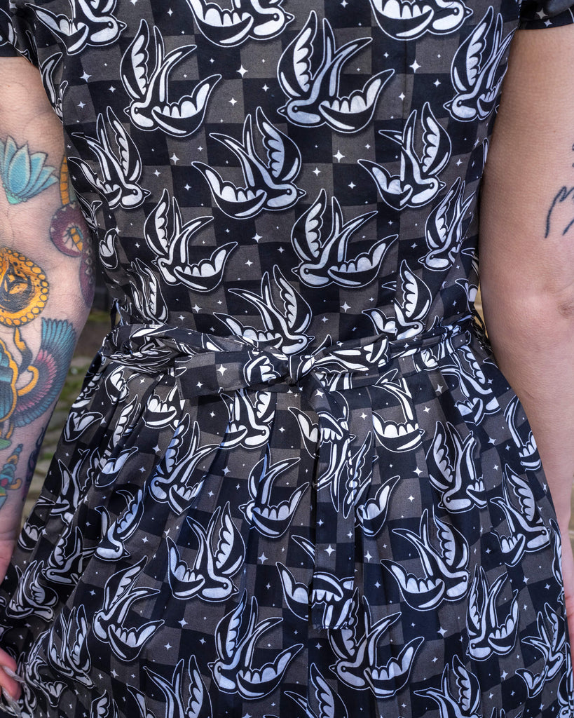 Close-up of the Run & Fly Swallow Print Dress, showing the black and white tattoo-inspired swallow design with a subtle checkered background. The fabric belt cinches the waist for a flattering fit. Run & Fly is a small ethical slow fashion company known for bold prints and comfy styles.