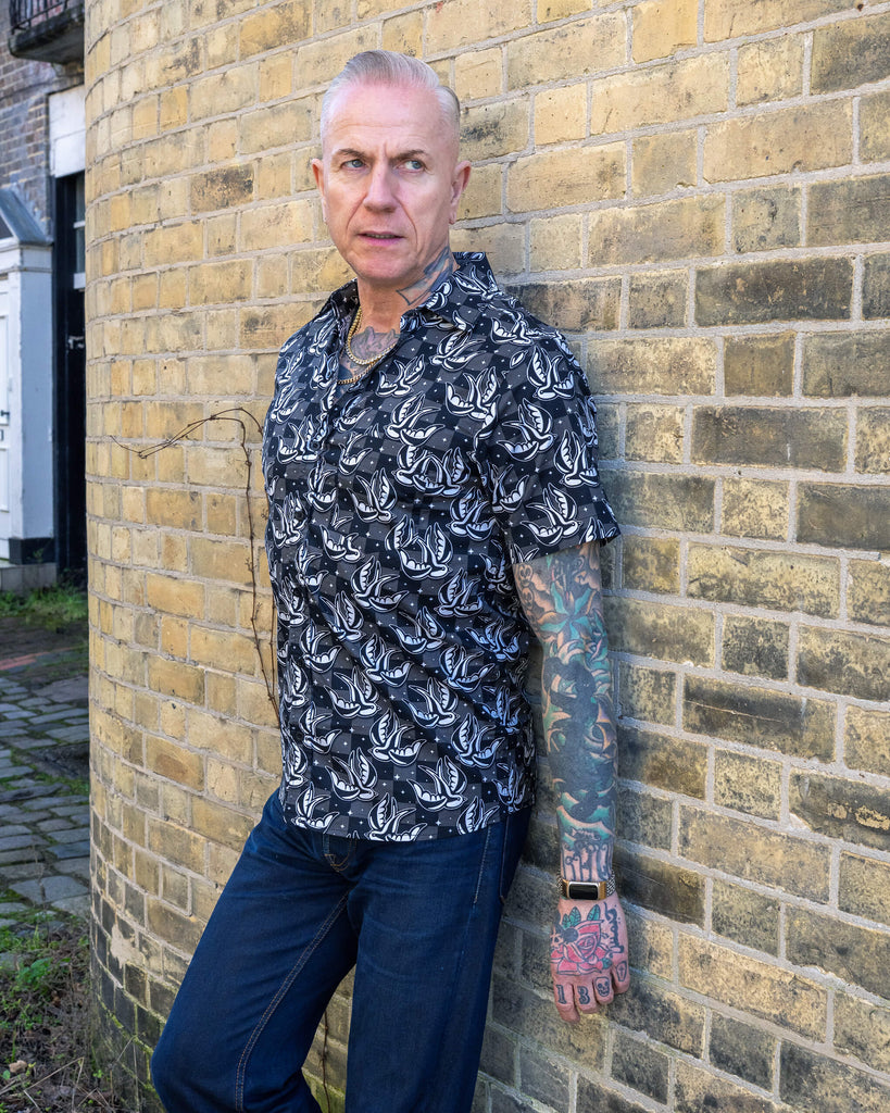 A stylish tattooed man models the Run & Fly swallow print short-sleeve shirt, posing casually against a curved brick wall. The black and white bird design stands out against the subtle check background, exemplifying Run & Fly's small-batch, slow fashion ethos.