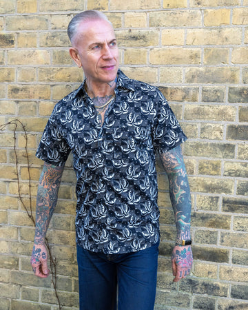 A tattooed male model with slicked-back grey hair wears the Run & Fly swallow print short-sleeve shirt while standing against a yellow brick wall. The relaxed fit and bold design reflect the brand's commitment to ethical slow fashion.
