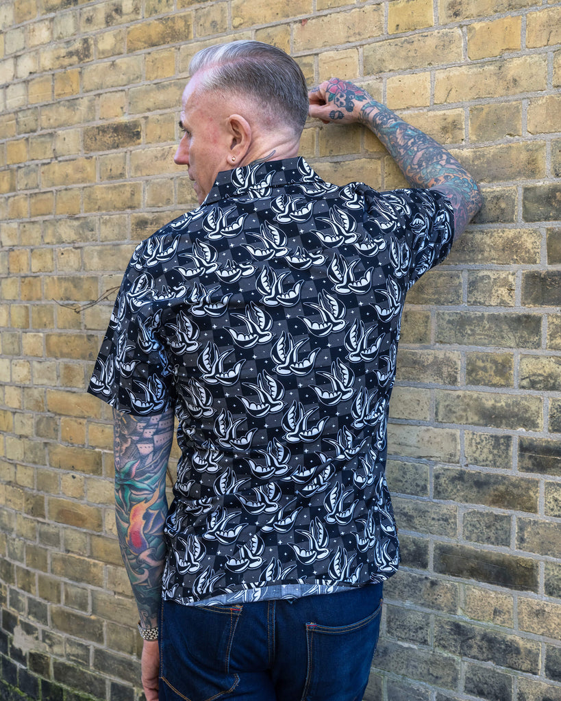 A tattooed man with short grey hair models a Run & Fly short-sleeve button-up shirt featuring a black and white swallow print on a subtle checkered background. He stands against a brick wall, showing the shirt's back detail. Run & Fly is a small ethical slow fashion company known for bold, unique designs.