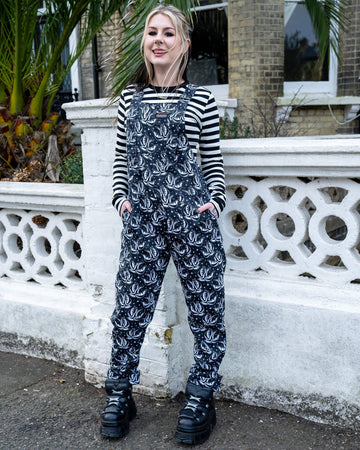 A person smiles while standing outdoors in Run & Fly dungarees with a black and white swallow print. They’re styled with a striped top and chunky platform boots in front of a white wall and palm tree.
