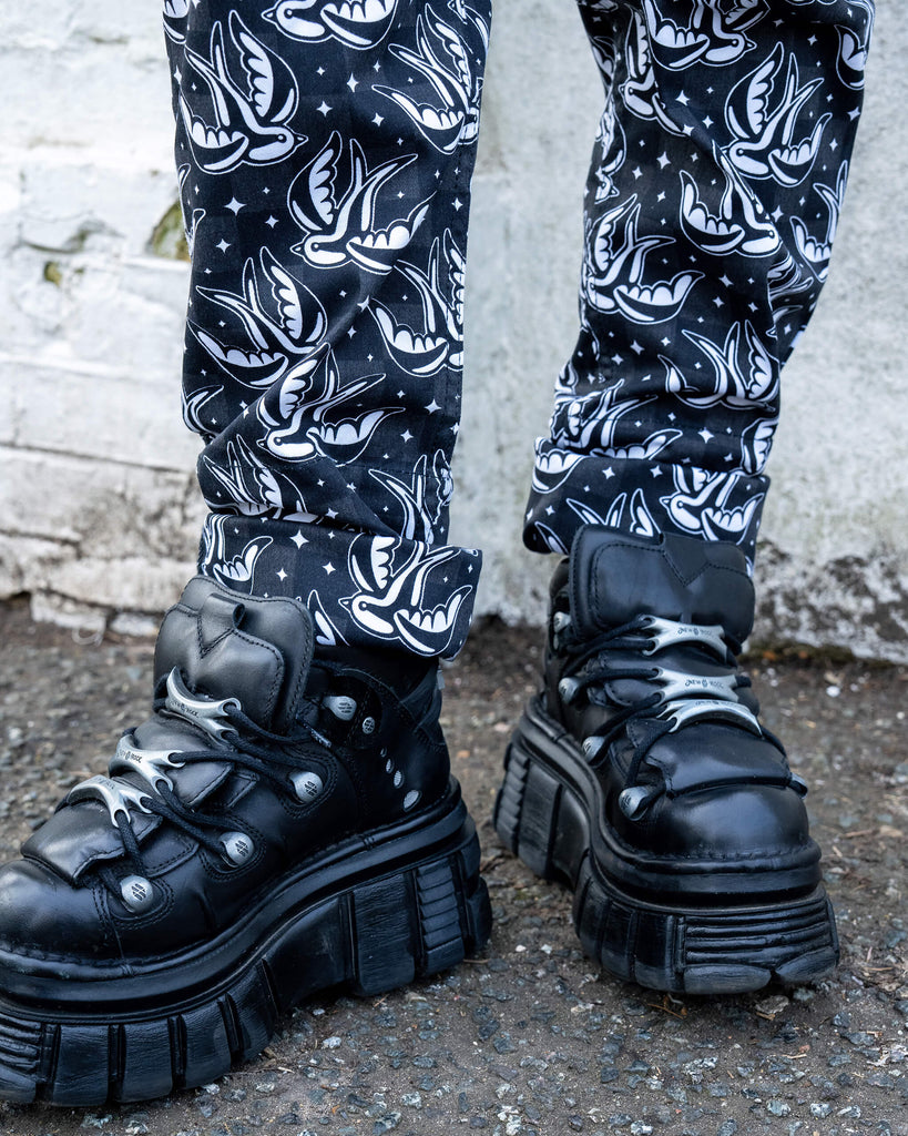 A close-up shot of black platform boots and the hem of the Run & Fly swallow print dungarees, rolled up at the ankles. The bold white swallow design pops against the black background.