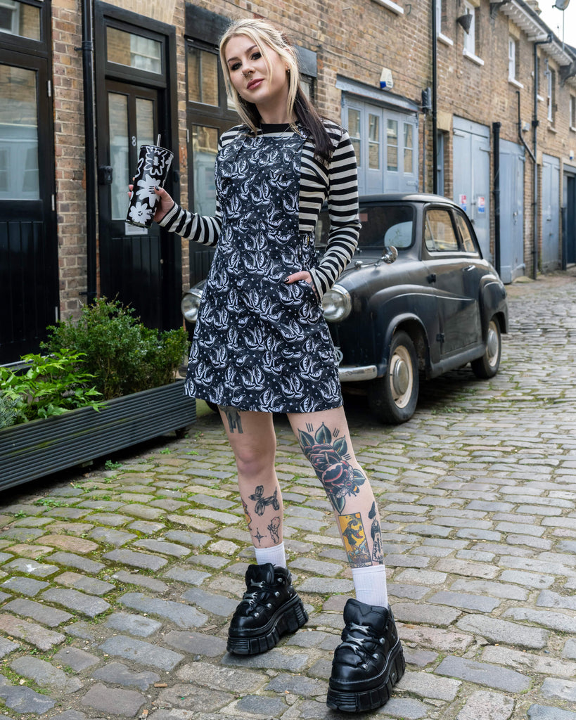 A person poses outdoors on cobblestones wearing a black and white Run & Fly swallow print pinafore dress, styled with striped sleeves, white socks, and platform boots. They hold a black floral reusable cup and show off colourful tattoos on their legs.