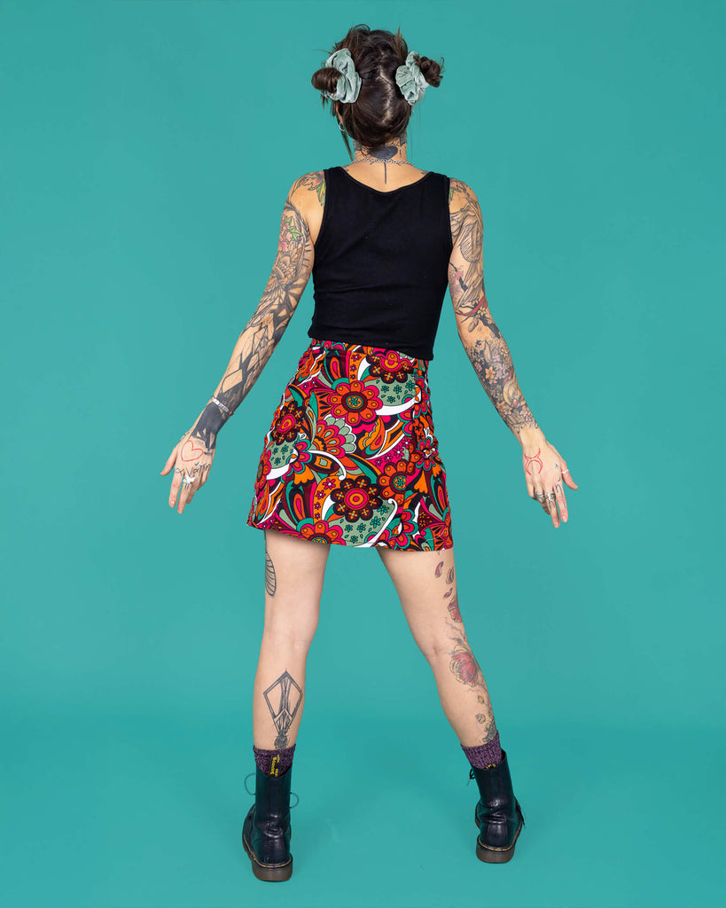 Yo a tattooed femme model is wearing a black vest and swirly floral A line skirt in a studio against a green background. They are facing the back