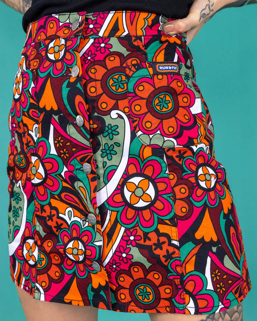 close up of the skirt worn by Yo a tattooed femme model is wearing a black vest and swirly floral A line skirt in a studio against a green background