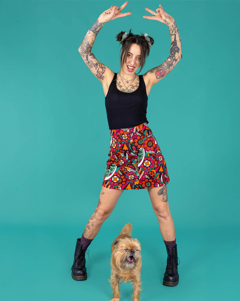Yo a tattooed femme model is wearing a black vest and swirly floral A line skirt in a studio against a green background