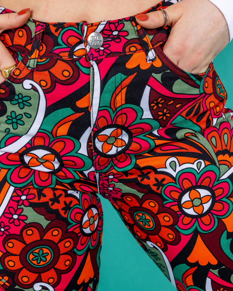 close up of the 70's style swirly floral print trousers and the rUN & fly logo button on a green background
