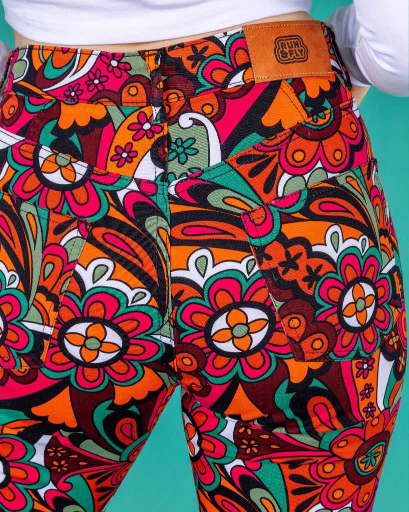 Amy a blonde model with bobbed hair is wearing a white long sleeved cropped tee with jazzy 70's swirly flares with black boots on a green background close up of the back