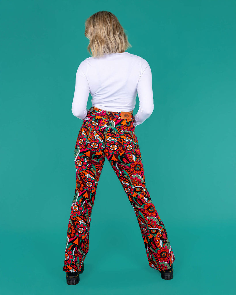 Amy a blonde model with bobbed hair is wearing a white long sleeved cropped tee with jazzy 70's swirly flares with black boots on a green background she is facing the back