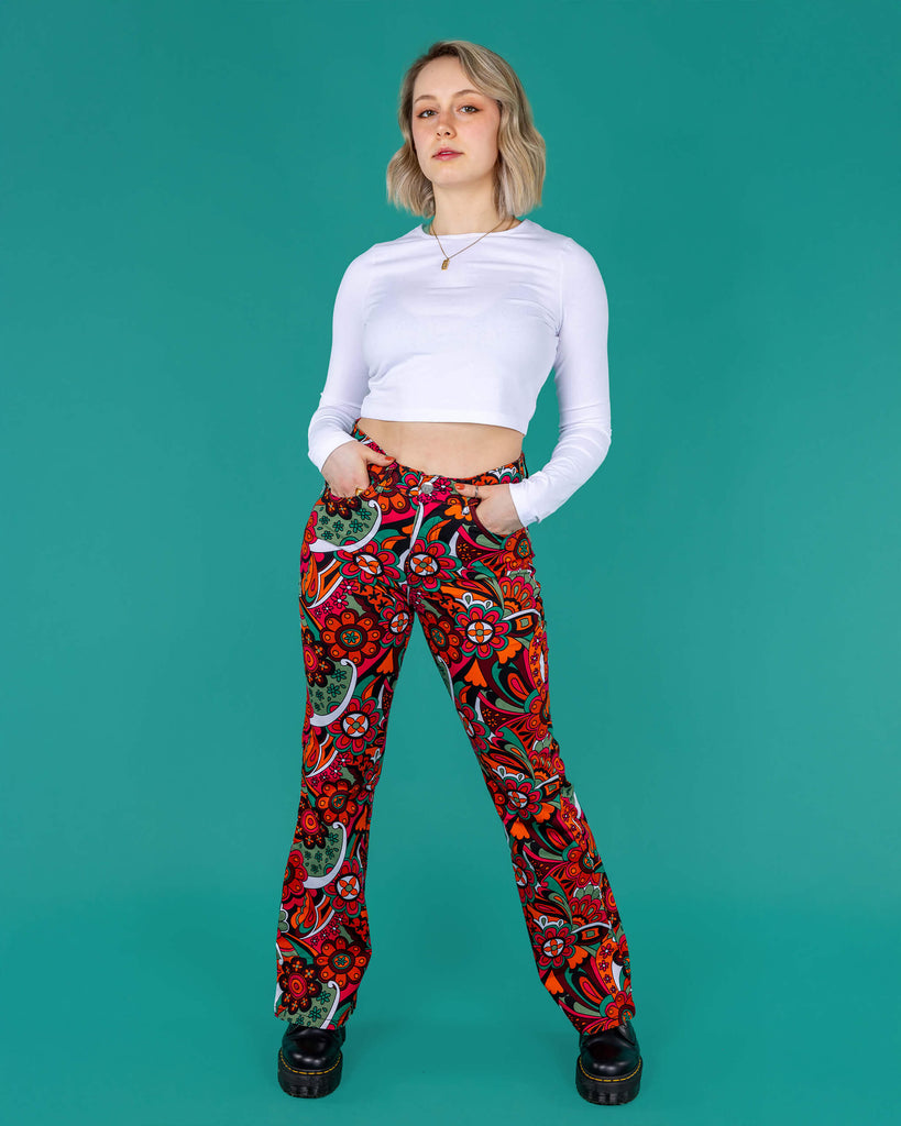 Amy a blonde model with bobbed hair is wearing a white long sleeved cropped tee with jazzy 70's swirly flares with black boots on a green background 