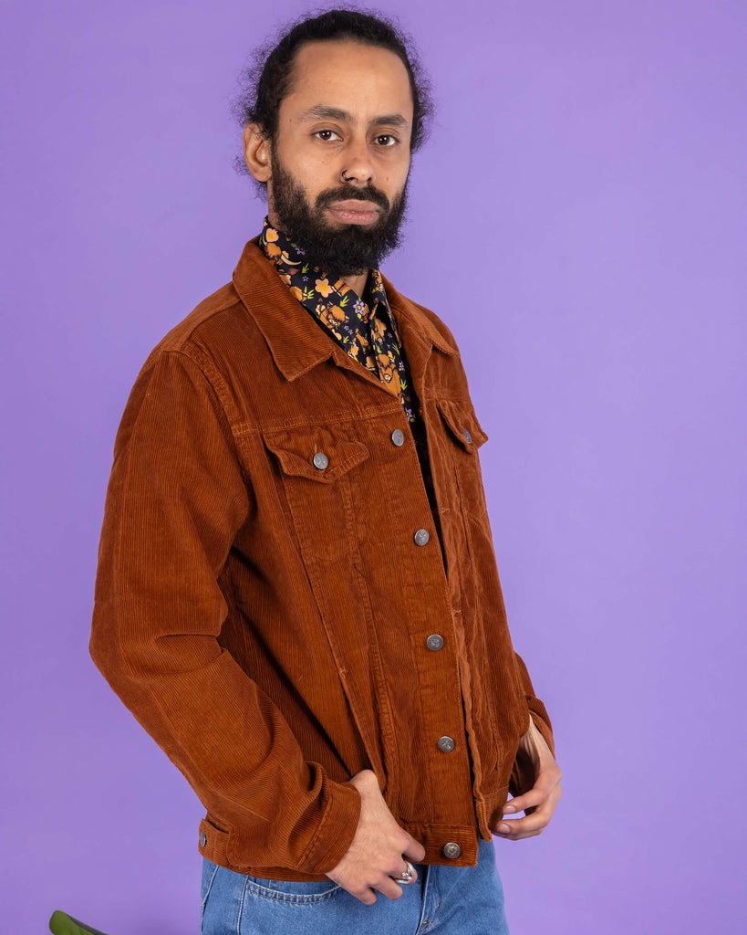 Richard, a hispanic male model with dark hair in a bun and a beard, is stood in a photography studio in Hove in front of a lilac backdrop wearing Retro Vintage Tan Corduroy Unisex Western Jacket with Highland Cows Short Sleeve Shirt underneath and blue jeans. Richard is stood slightly to the side posing to the camera with his thumbs resting in the jacket pockets. The photo is cropped at the thighs.