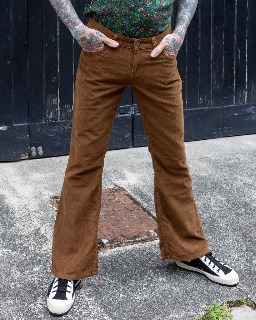 James is stood in front of a black door background wearing the tobacco cord bootcut flares with green paisley shirt. They are facing forward with both hands in the pockets of the flares