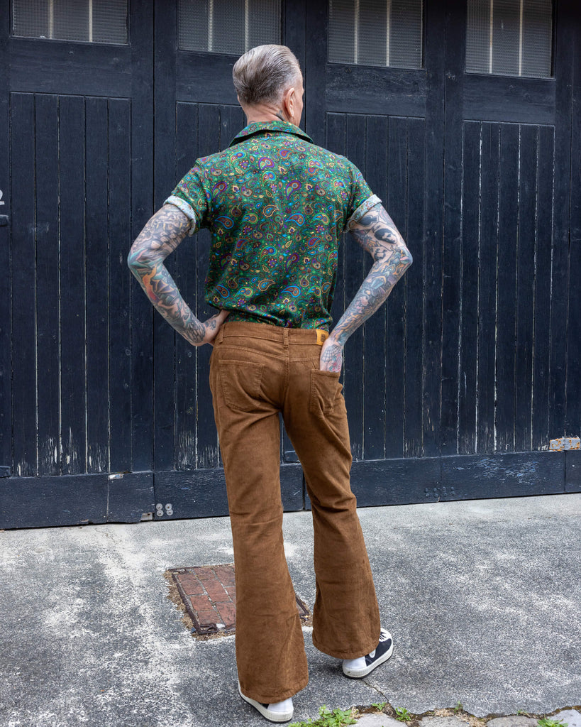James, a tattooed male model, is stood outside in Hove in a mews wearing Tan Corduroy Bootcut Flares with Forest Green Paisley Short Sleeve Rayon Shirt. James is stood with his back to the camera with one leg in front of the other. He has one hand on his hip and the other in the back pocket of the trousers.
