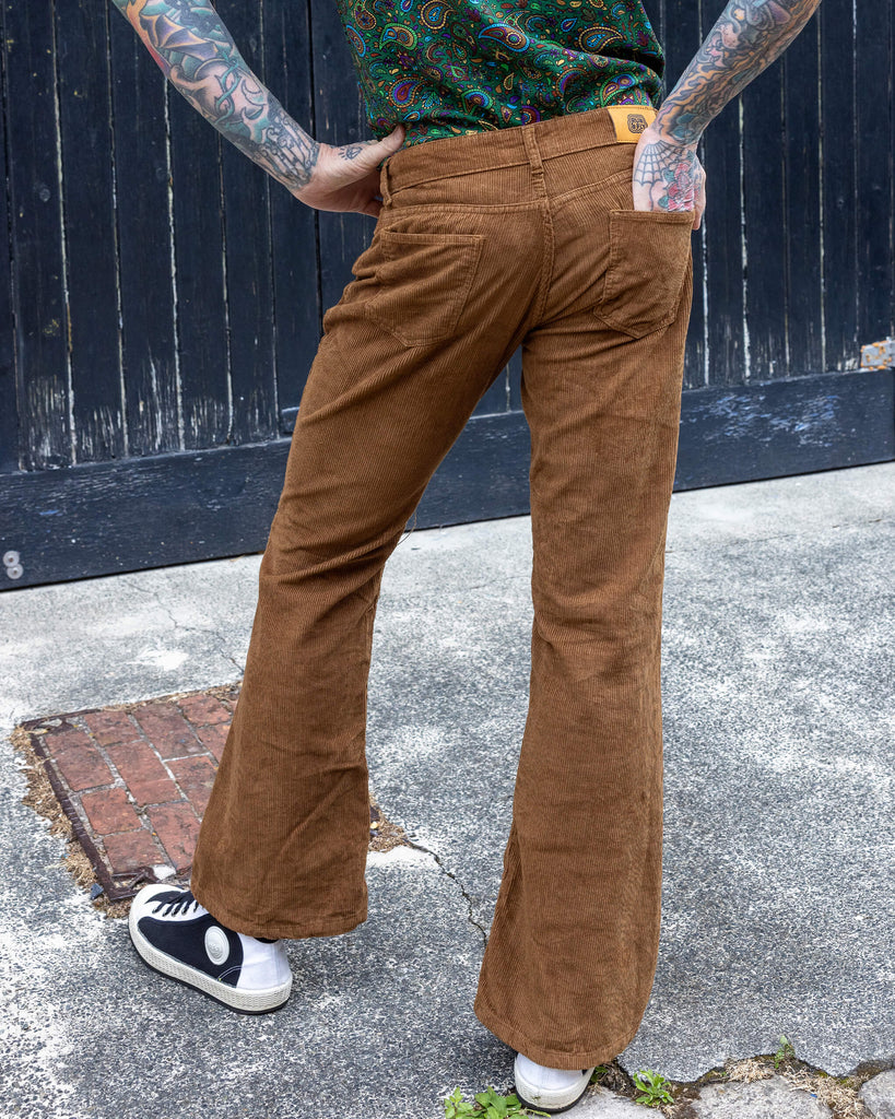 James, a tattooed male model, is stood outside in Hove in a mews wearing Tan Corduroy Bootcut Flares with Forest Green Paisley Short Sleeve Rayon Shirt. James is stood with his back to the camera with one leg in front of the other. He has one hand on his hip and the other in the back pocket of the trousers.