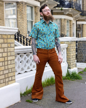 Dec is stood outside a town house in Hove wearing the Tobacco Corduroy Bell Bottom Super Flares Trousers with Bunny Meadow Short Sleeve Shirt. He is leaning against a wall with one leg bent and his hand on his hip. He is smiling looking off into the distance.