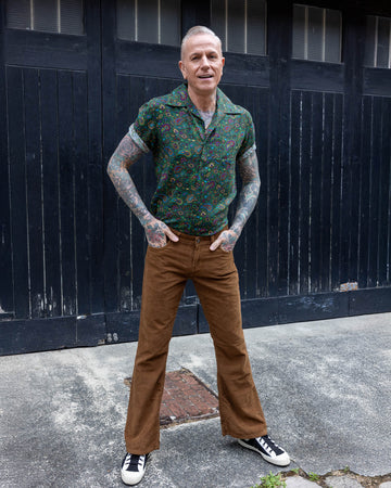 James, a tattooed male model, is stood outside in Hove in a mews wearing Tan Corduroy Bootcut Flares with Forest Green Paisley Short Sleeve Rayon Shirt. James is stood facing the camera with his hands in his pockets and looking off to one side smiling. 