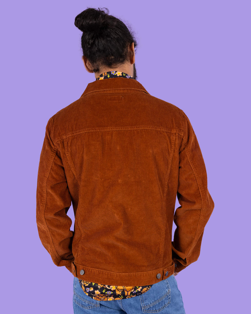 Richard, a hispanic male model with dark hair in a bun and a beard, is stood in a photography studio in Hove in front of a lilac backdrop wearing Retro Vintage Tan Corduroy Unisex Western Jacket with Highland Cows Short Sleeve Shirt underneath and blue jeans. Richard is posing with his back to the camera and arms in front of his. The photo is cropped at the thighs.