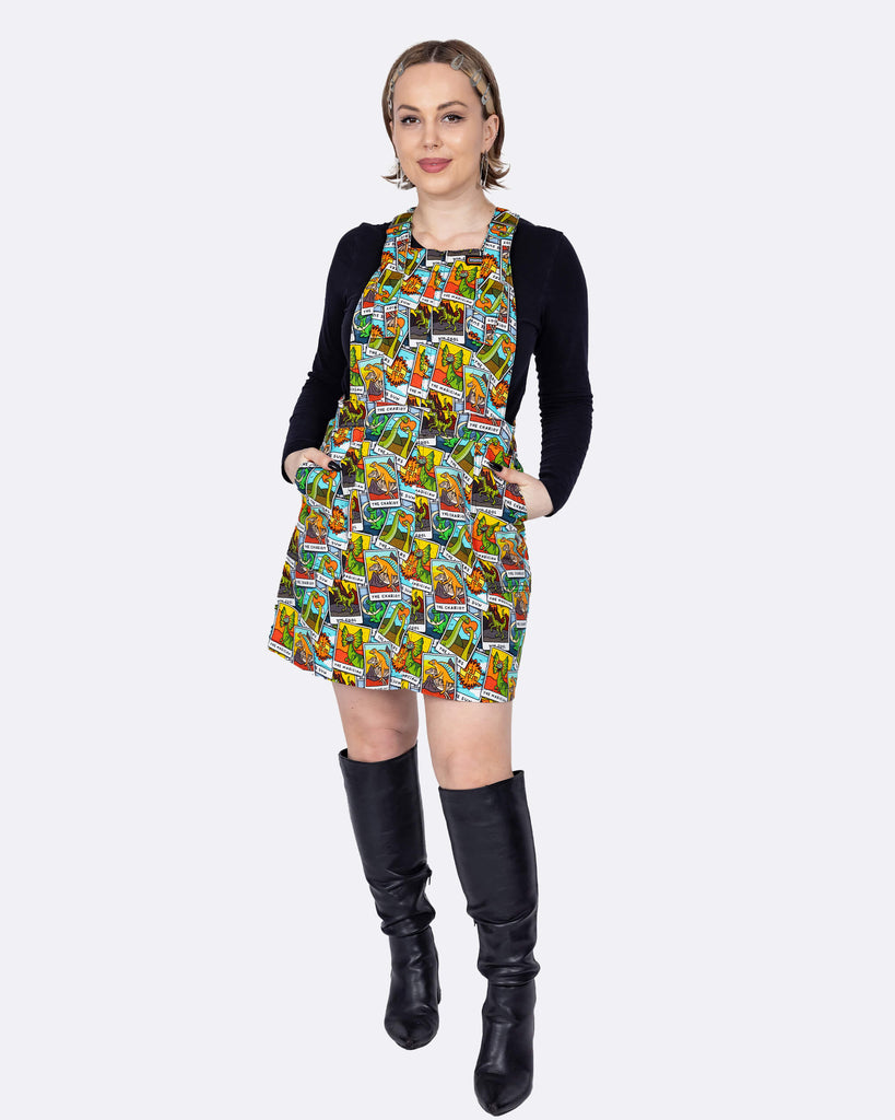 The Tarot Dinosaurs Stretch Twill Pinafore Dress worn by a femme model with short blonde hair with a black long sleeve top and black knee high boots on a white background. She is facing forward smiling leaning more on one leg with both hands resting in the pinafore pockets. The mini pinafore print has an aqua blue base with clouds with various scattered tarot cards with dinosaurs.