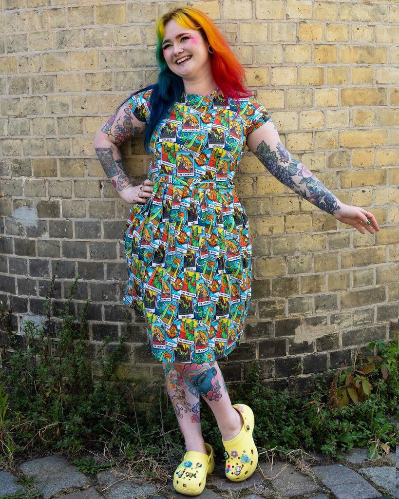 A woman with rainbow-coloured hair and vibrant makeup standing and smiling looking off to one side, wearing Tarot Dino Stretch Belted Tea Dress with Pockets. The dress has an all over colourful dinosaur tarot card print. She has colourful tattoos and is wearing yellow Crocs with funl pins. She is posing with one hand on her hip and other stretched out to her side against a yellow brick wall. The dress is from the ethical brand Run & Fly.