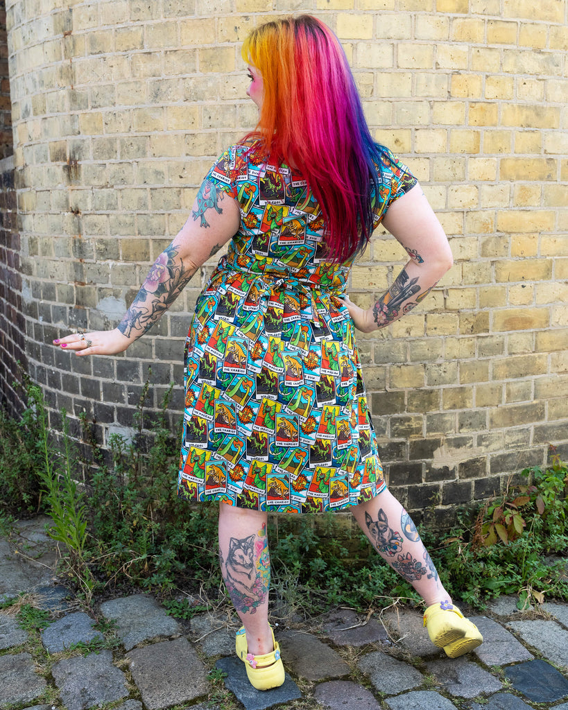 A woman with rainbow-coloured hair and vibrant makeup standing and smiling looking off to one side, wearing Tarot Dino Stretch Belted Tea Dress with Pockets. The dress has an all over colourful dinosaur tarot card print. She has colourful tattoos and is wearing yellow Crocs with funl pins. She is posing with one hand on her hip and other stretched out to her side against a yellow brick wall. The dress is from the ethical brand Run & Fly.