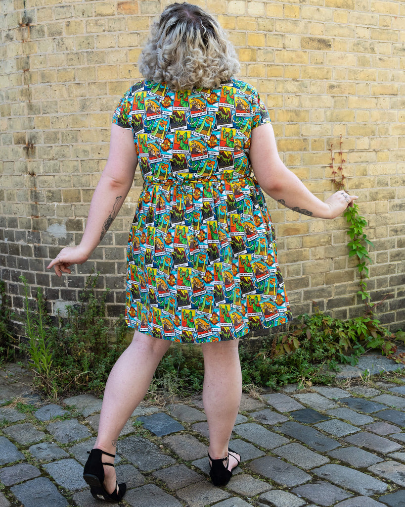 Rear view of a woman with curly blonde hair, showcasing the back of the Tarot Dino Stretch Belted Tea Dress with Pockets. The dress has an all over colourful dinosaur tarot card print. She has tattoos on her arms and is standing in front of a yellow brick wall. The dress is designed by the ethical, slow fashion brand Run & Fly.