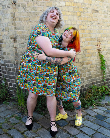Two women, one with curly blonde hair and the other with rainbow-coloured hair, hugging and laughing. They are both wearing Tarot Dino Stretch Belted Tea Dress with Pockets. The dress has an all over colourful dinosaur tarot card print. They are standing against a yellow brick wall. The dresses are made by the sustainable fashion brand Run & Fly.
