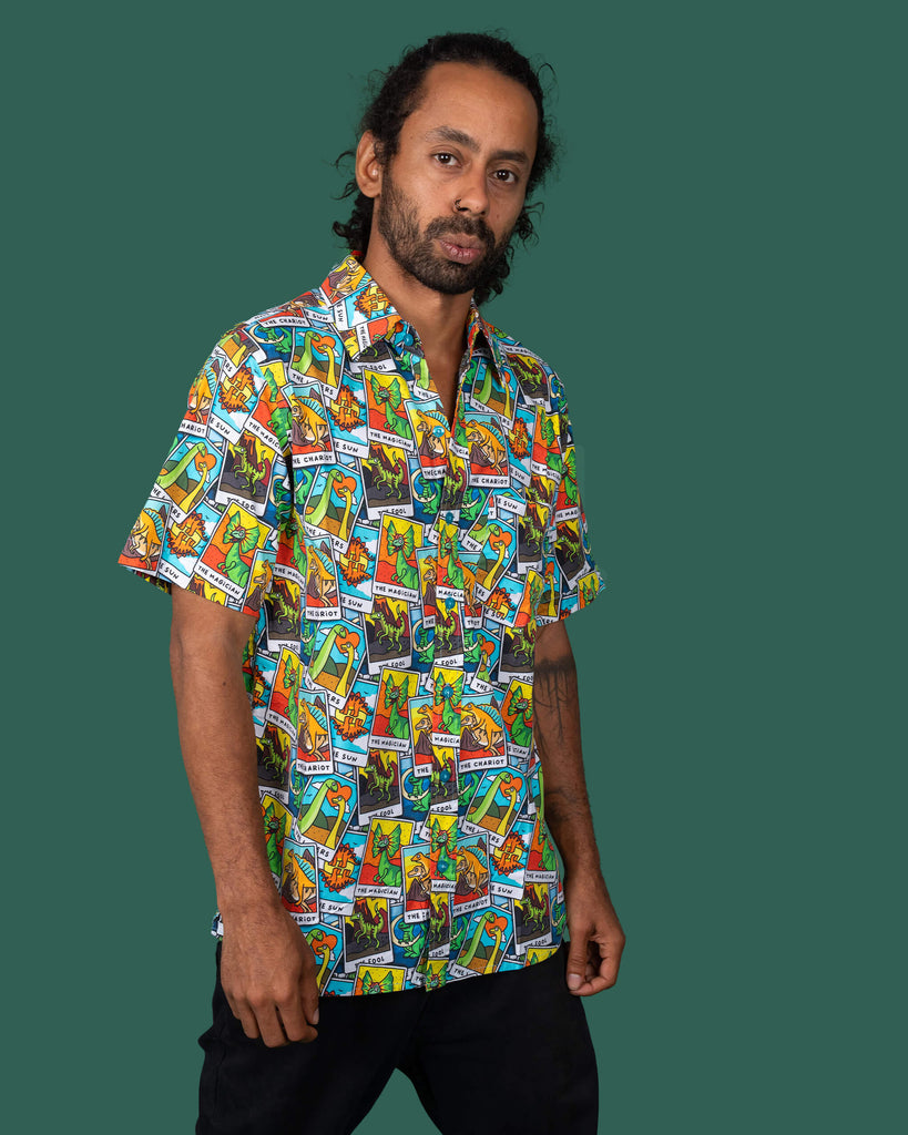 Model posing with hands in pockets, wearing a Run & Fly short-sleeve shirt featuring a whimsical dinosaur tarot card pattern. The outfit includes black trousers, showcasing the brand's fun and sustainable fashion approach.
