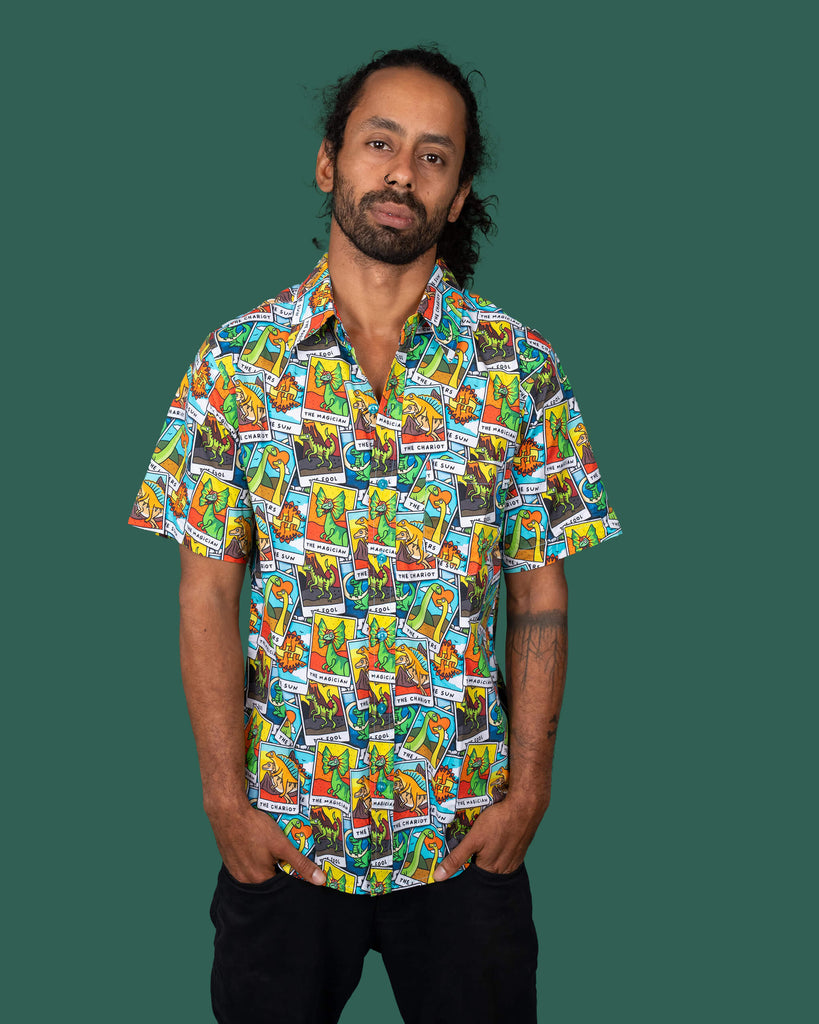 Model posing with hands in pockets, wearing a Run & Fly short-sleeve shirt featuring a whimsical dinosaur tarot card pattern. The outfit includes black trousers, showcasing the brand's fun and sustainable fashion approach.