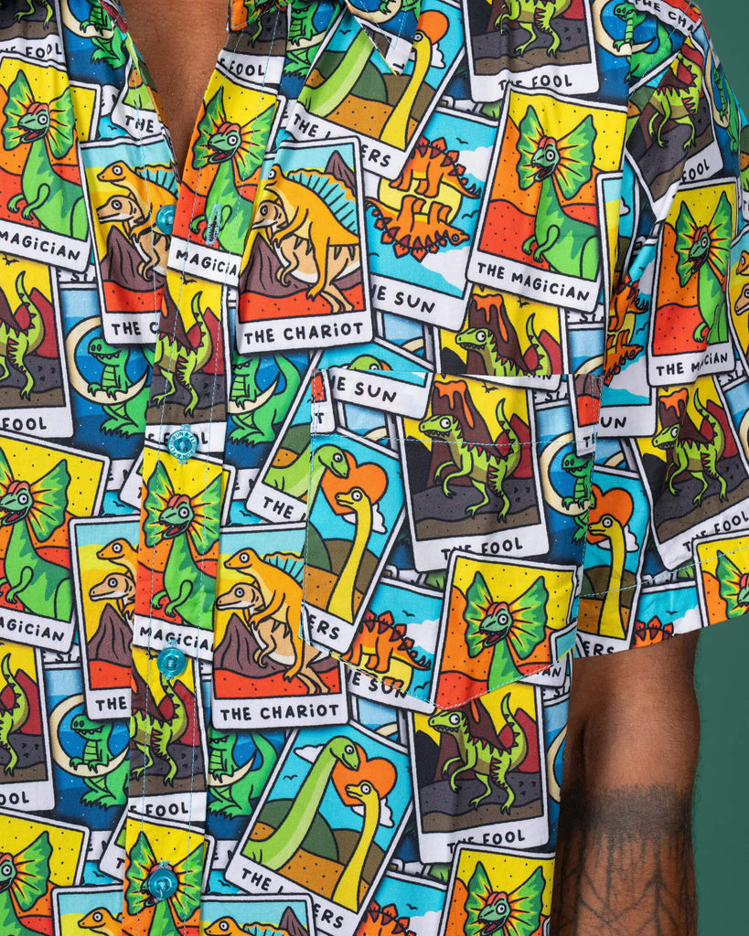 Close-up view of a Run & Fly short-sleeve shirt featuring a colourful dinosaur tarot card pattern. The detailed design includes various dinosaurs depicted as tarot card figures like The Magician, The Chariot, and The Sun, highlighting the brand's fun and ethical slow fashion approach.