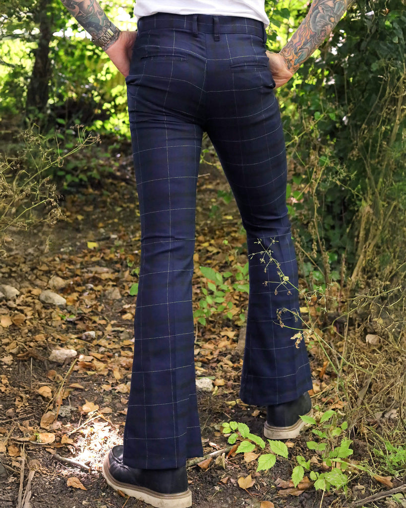 Model is stood outside in a wooded area wearing the blue tartan stretch plaid bell bottom trousers in dark blue. He is heavily tattooed and paired the trousers with a white t-shirt and brown boat shoes. He is posing facing away from the camera with both hands in his pockets. Photo is cropped from the hips down.
