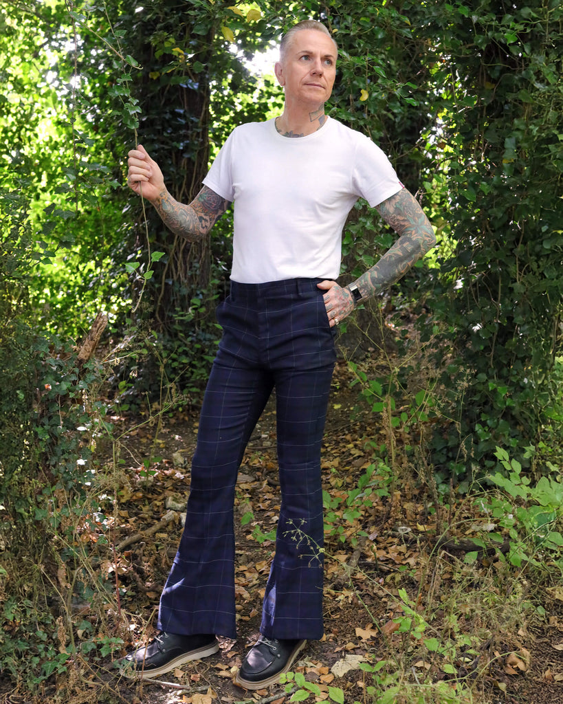 Model is stood outside in a wooded area wearing the blue tartan stretch plaid bell bottom trousers in dark blue. Model is posing facing towards the camera with one leg to the side, one hand is in his pocket whilst the other is resting by his side. Photo is cropped from the waist down. He has paired the trousers with a white t-shirt and brown boat shoes.