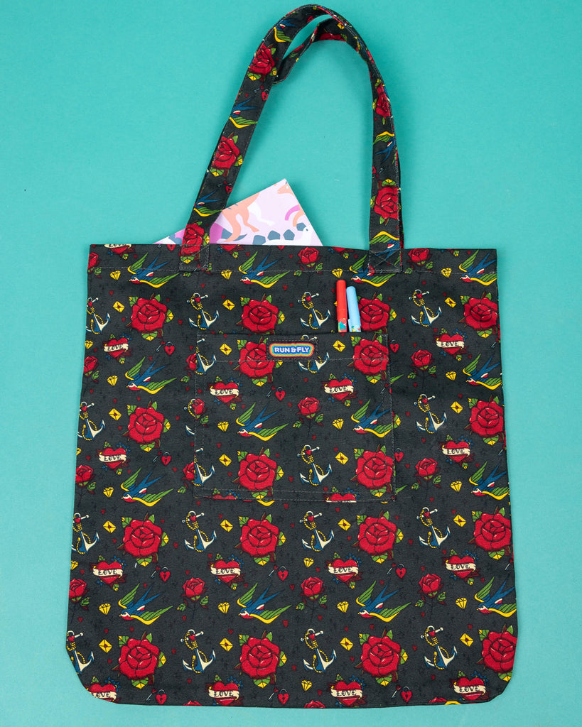 a dark green tote bag with all over tattoo flash style design including red roses, swallows, anchors, love hearts. The tote bag is laid flat on a green background with a book in the main section of the bag and two pens in the smaller front pocket with has Run & Fly logo on the front. 