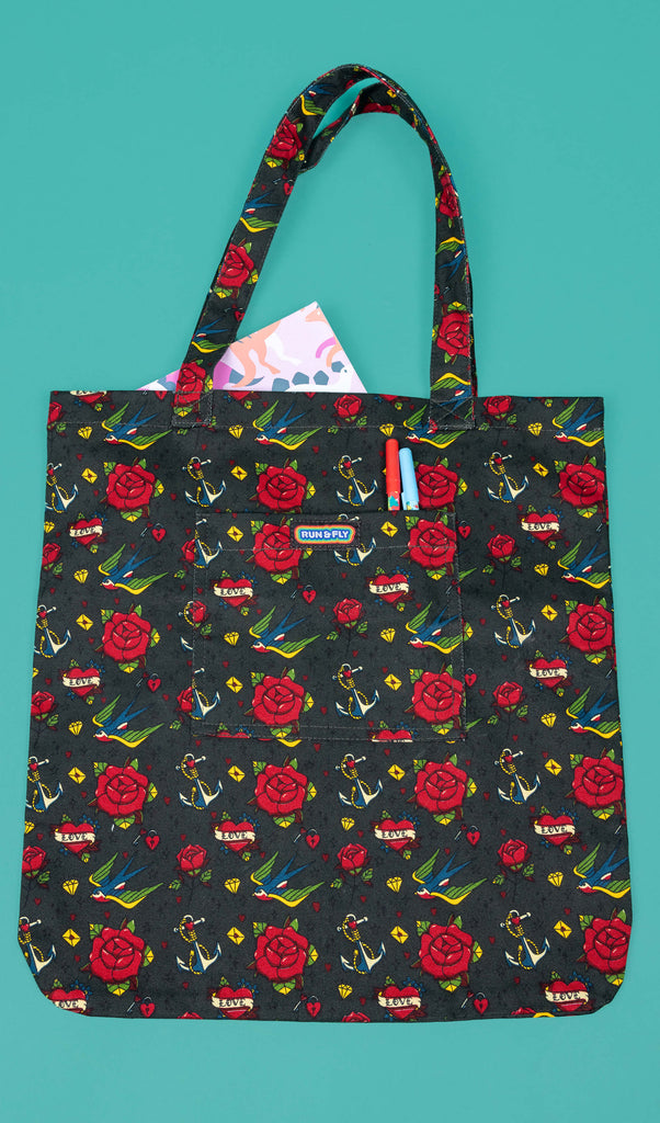 a dark green tote bag with all over tattoo flash style design including red roses, swallows, anchors, love hearts. The tote bag is laid flat on a green background with a book in the main section of the bag and two pens in the smaller front pocket with has Run & Fly logo on the front. 