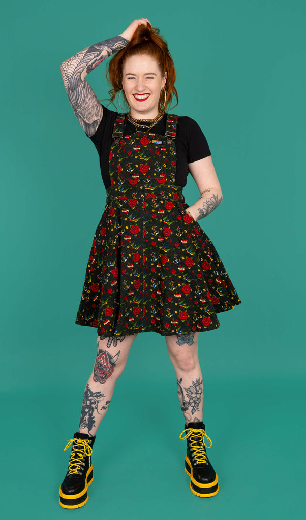 A tattooed femme model with red hair wearing the run and fly retro old school tattoo flared pinafore dress with yellow and black platform boots, facing forward smiling with one hand in the dress pocket whilst the other scrunches their hair. The print has traditional tattoo style red roses, blue swallows, yellow envelopes, cream anchors and valentines love heart. The flared pinafore features a run and fly rainbow tag on the front bib pocket and has adjustable clip straps with Run & Fly logo.