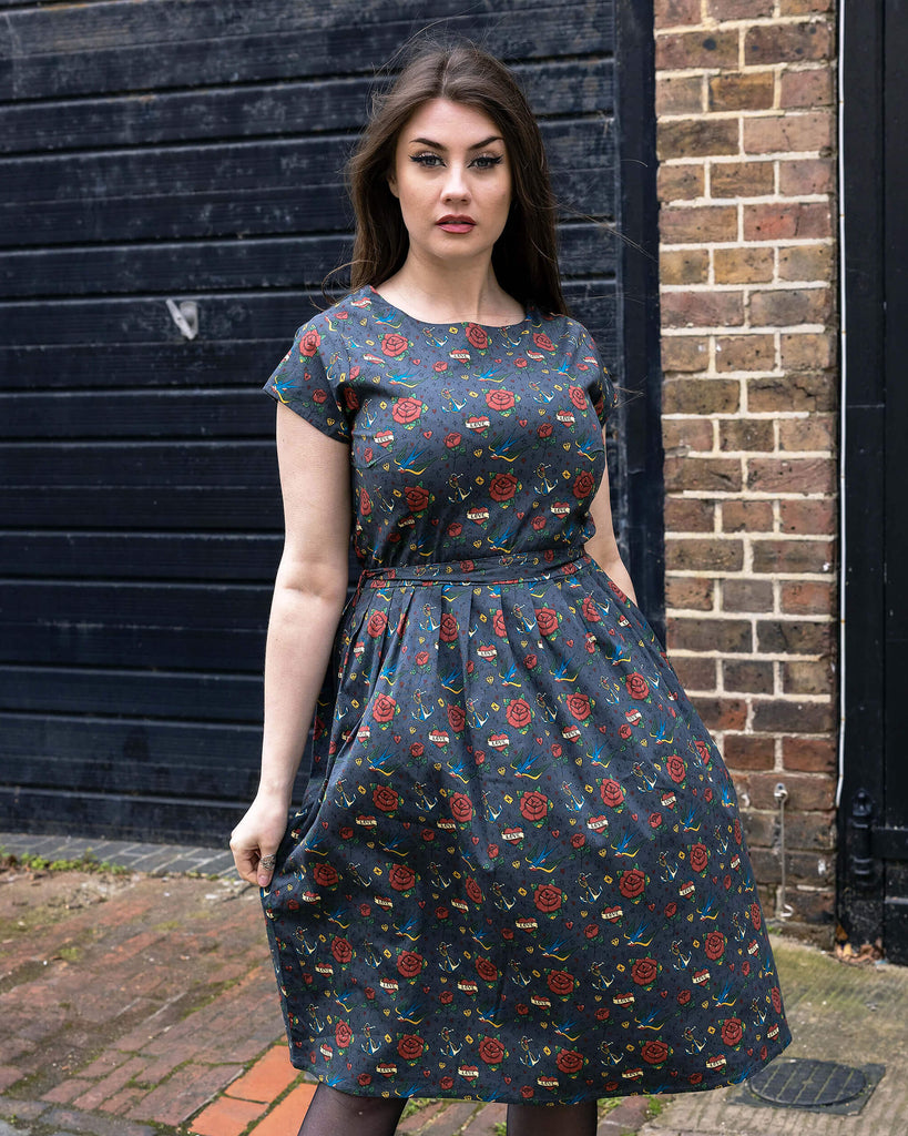 Charlotte is wearing the Run & Fly Retro Tattoo Stretch Belted Tea dress with pockets, grey with red roses, hearts and birds print, and waist belt. Model is looking at the camera, and standing in a cobbled street in front of garage doors.