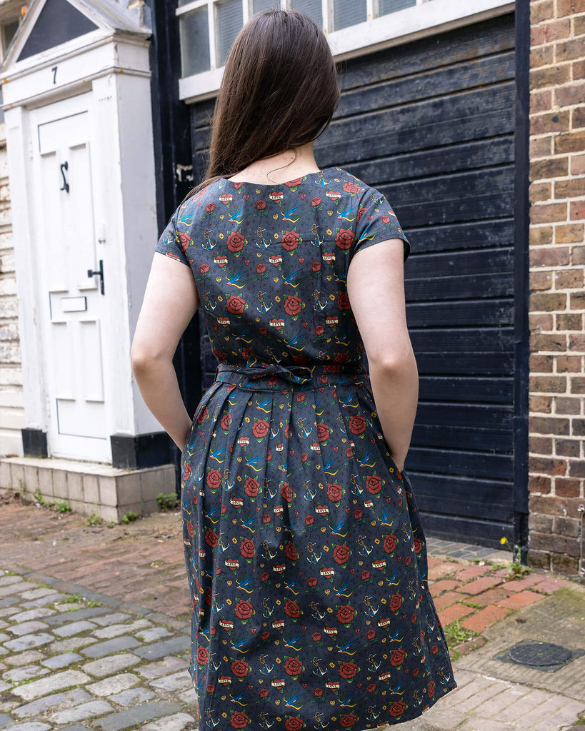 Charlotte is wearing the Run & Fly Retro Tattoo Stretch Belted Tea dress with pockets, grey with red roses, hearts and birds print, and waist belt. Model has her back to the camera, with hands in side pockets and standing in a cobbled street in front of garage doors.