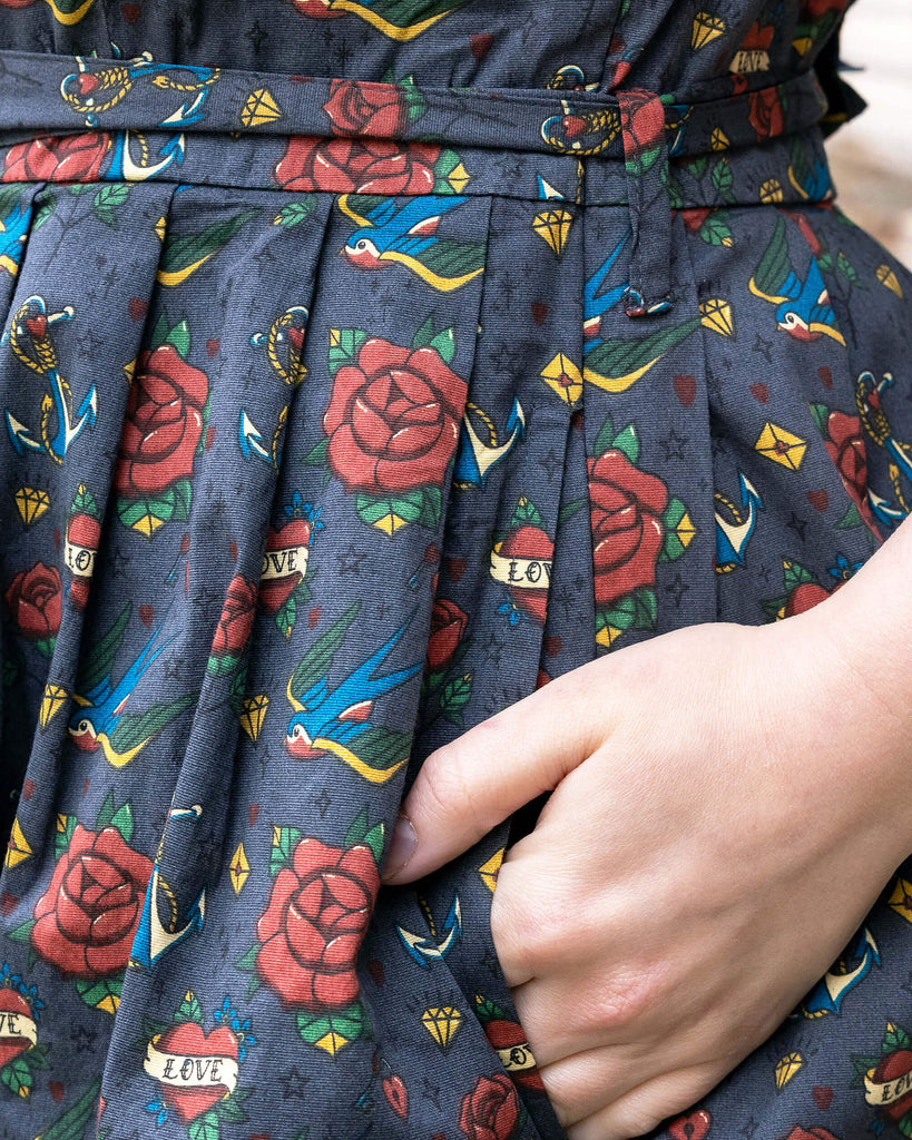 Closeup of the Run & Fly Retro Tattoo Stretch Belted Tea dress with pockets, grey with red roses, hearts and birds print, and waist belt. Model has hand in side pocket.