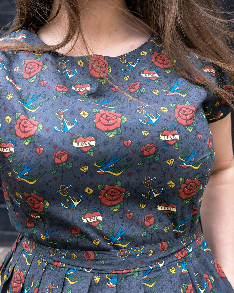 Closeup of the Run & Fly Retro Tattoo Stretch Belted Tea dress with pockets, grey with red roses, hearts and birds print, and waist belt. Model has hand in side pocket.