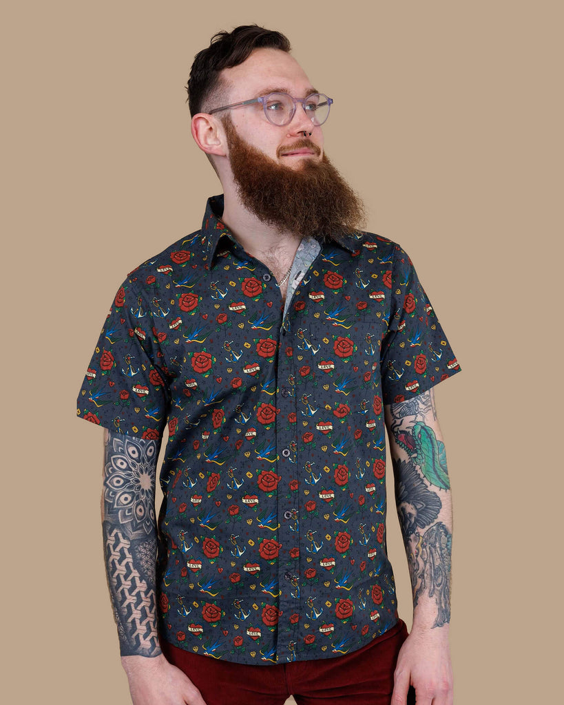 Dec, a white male model with a beard and glasses is wearing Retro Old School Tattoo Print Short Sleeve Shirt with Tawney Port Corduroy Bell Bottom Flares. The print has traditional tattoo style red roses, blue swallows, yellow envelopes, anchors and valentines love heart. Dec is stood in front of a beige backdrop at a photography studio in Hove posing facing the camera and looking off to one side with his arms by his sides. 